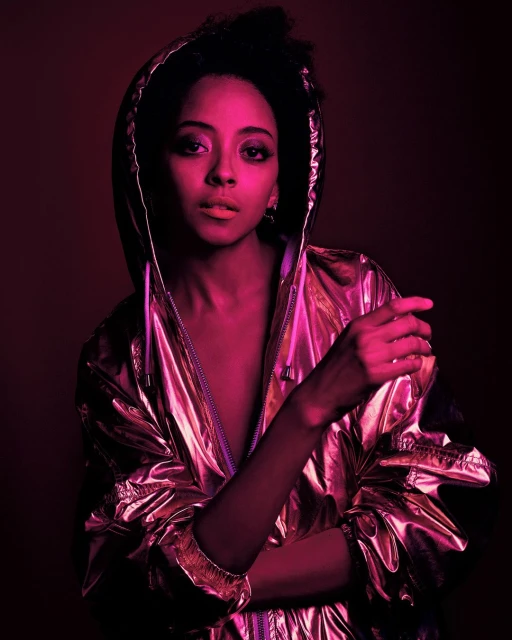 Close-up portrait of a Black female model wearing a metallic hooded jacket with arms crossed against a plain dark background.