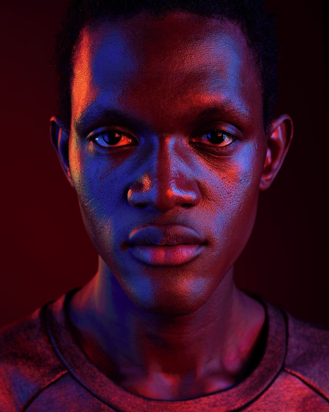 Portrait of a young Black man with short hair, looking directly at the camera, under dramatic red and blue lighting in a dark studio.
