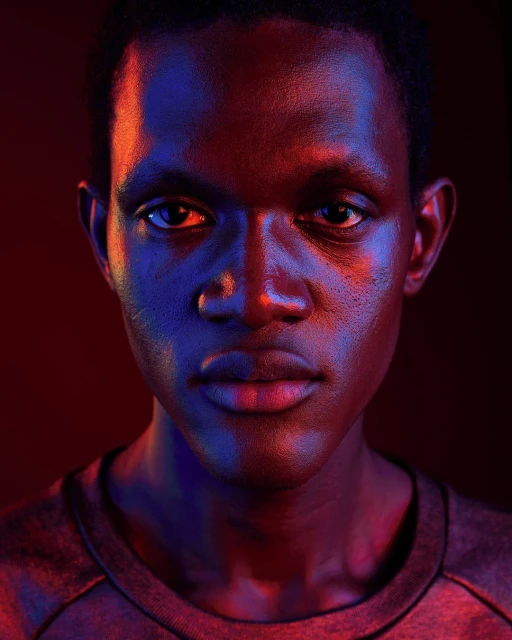 Portrait of a young Black man with short hair, looking directly at the camera, under dramatic red and blue lighting in a dark studio.