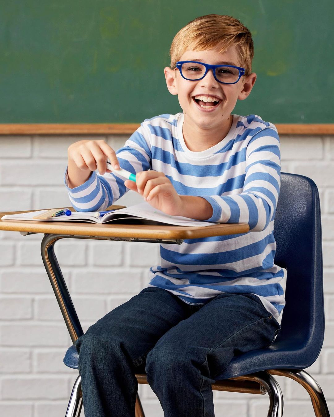 A smiling child with glasses sits at a school desk, holding a pen, with a textbook and green chalkboard in the background.