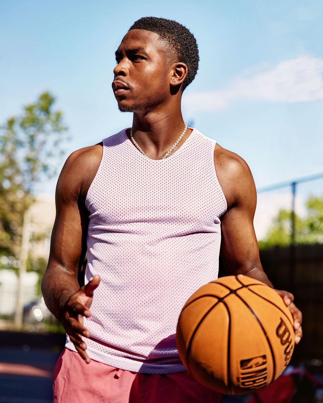 Young man in a tank top and shorts holding a basketball on an outdoor court. Bright daylight illuminates the sporty scene.