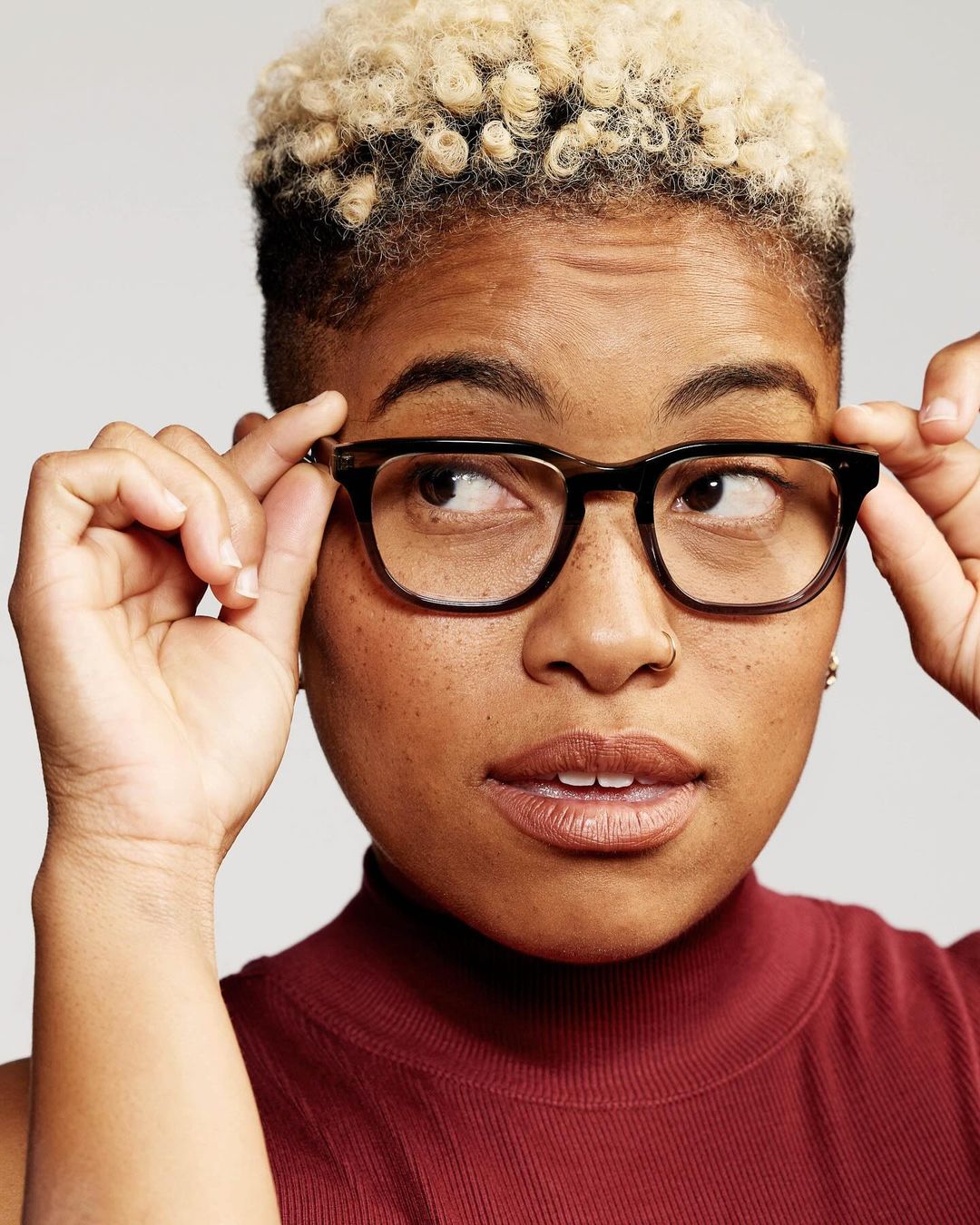 Close-up of a young adult female wearing glasses and a maroon top, looking to the side against a light gray background in a portrait shot.