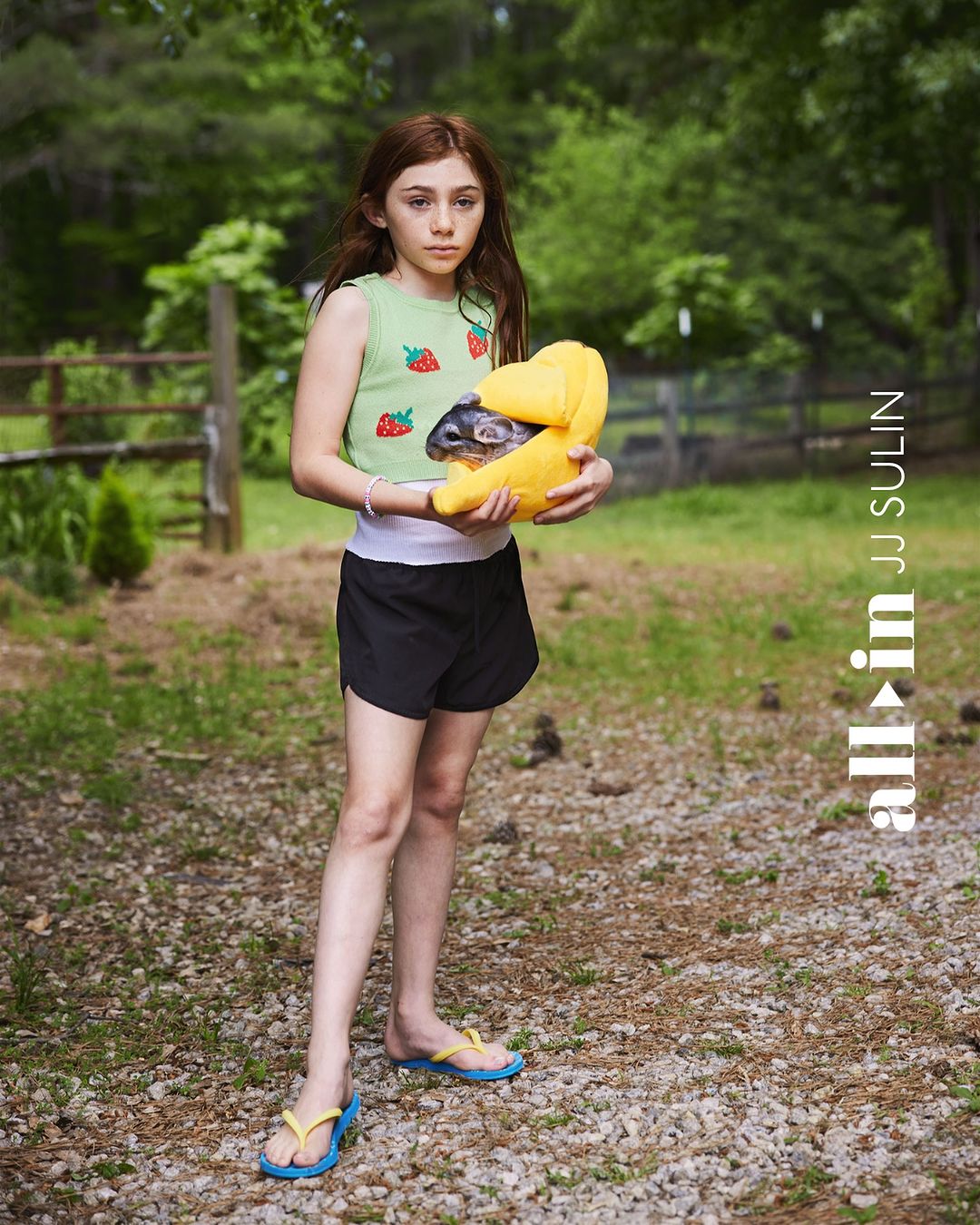 A young girl holding a chinchilla in a yellow carrier, standing outdoors in a rural setting with trees and a gravel path.