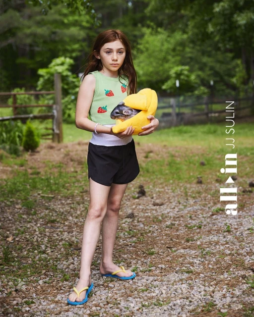 A young girl holding a chinchilla in a yellow carrier, standing outdoors in a rural setting with trees and a gravel path.
