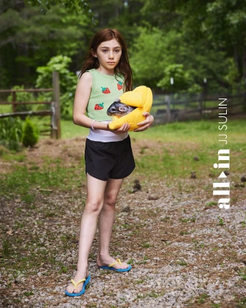 A young girl holding a chinchilla in a yellow carrier, standing outdoors in a rural setting with trees and a gravel path.