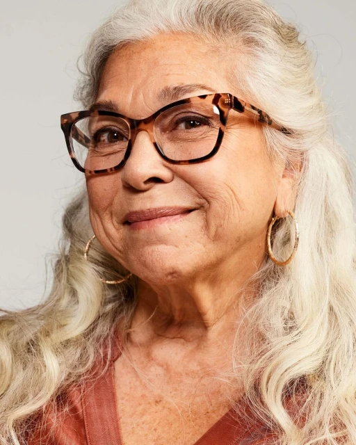 Close-up portrait of an elderly woman with gray hair wearing brown-framed glasses and gold hoop earrings against a neutral backdrop.