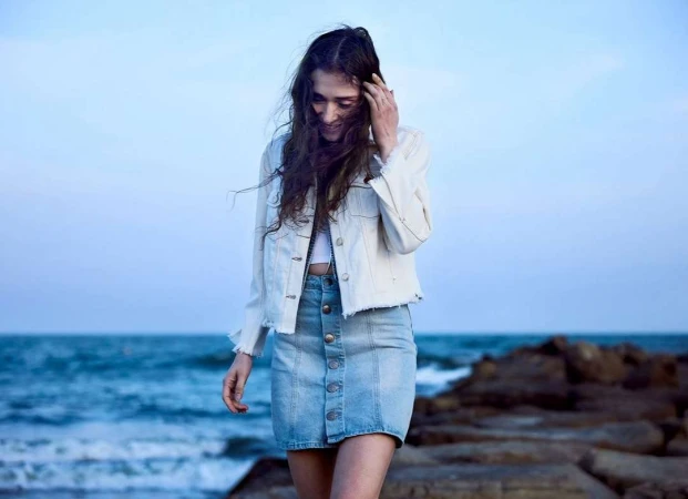 Woman wearing a white jacket and denim skirt walking near the ocean during dusk by photographer Jj Sulin.