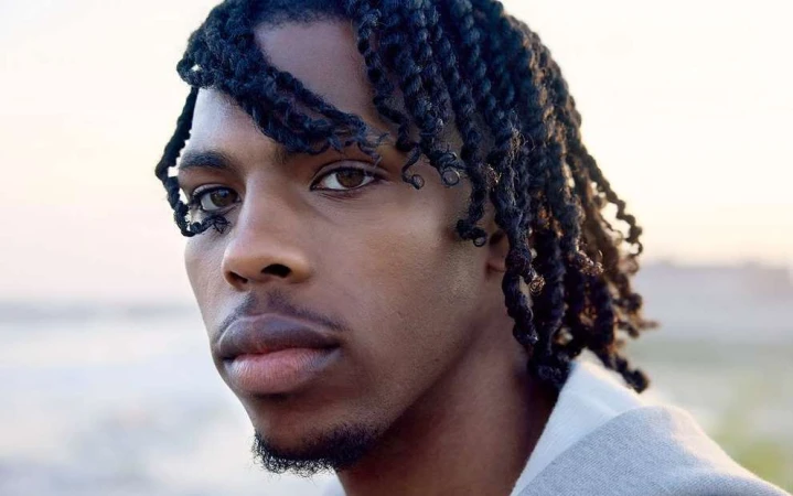 Close-up portrait of a young Black man with braided hair outdoors against a softly blurred horizon.