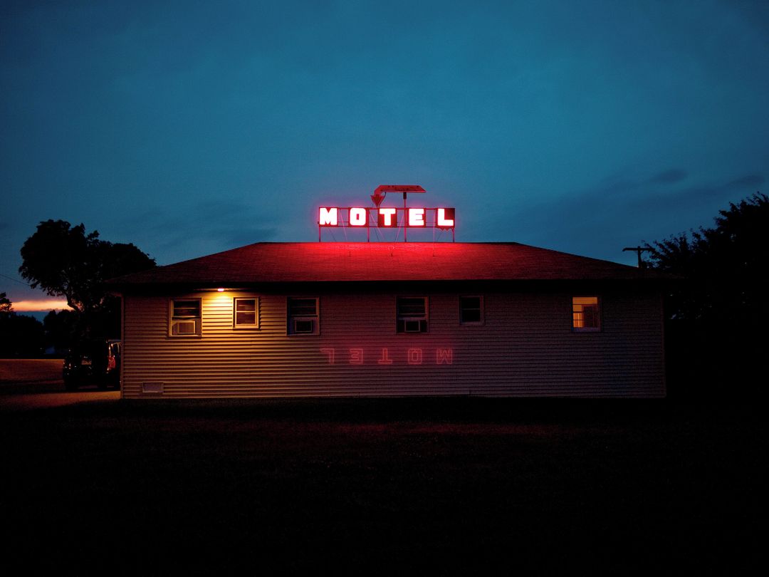 Motel exterior at twilight featuring a vintage neon sign on the roof, glowing against a dark blue sky. The sign's light reflects on the building.