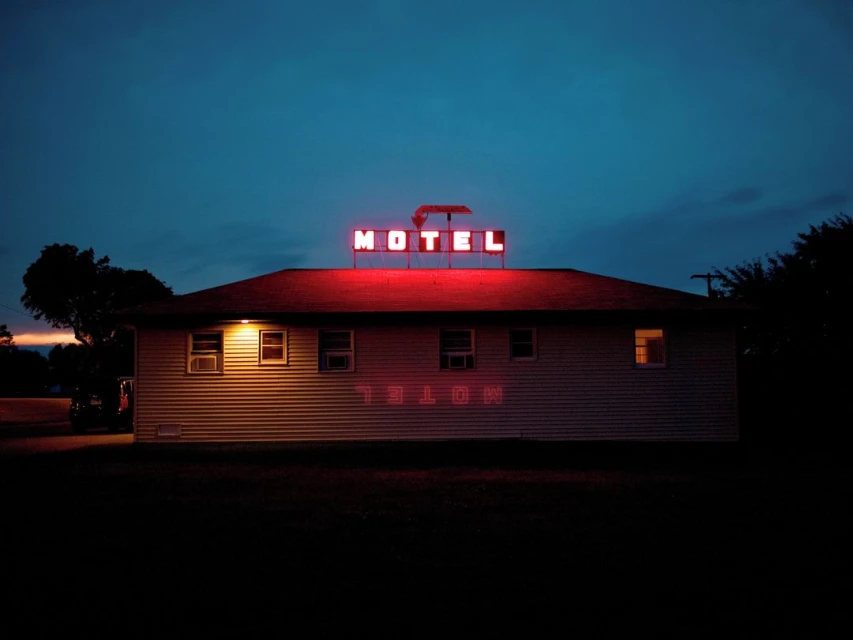 Motel exterior at twilight featuring a vintage neon sign on the roof, glowing against a dark blue sky. The sign's light reflects on the building.