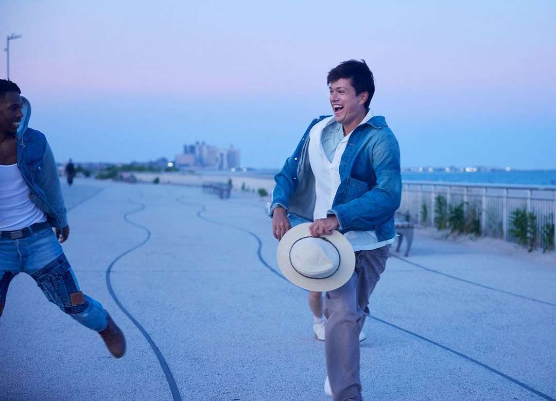 Two men in denim jackets stroll along a beachside walkway at dusk, one joyfully running with a hat in hand, with city buildings in the distance.