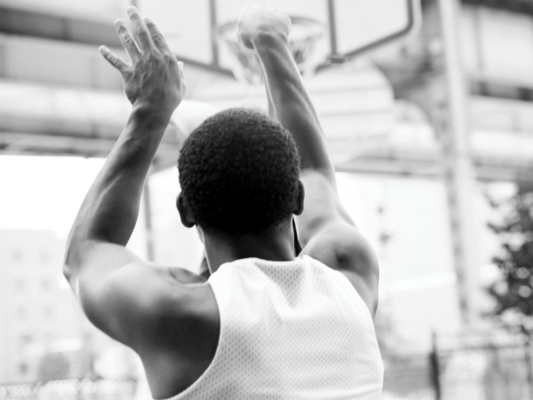 Black and white photo of an African American man in a white tank top shooting a basketball toward a hoop outdoors.