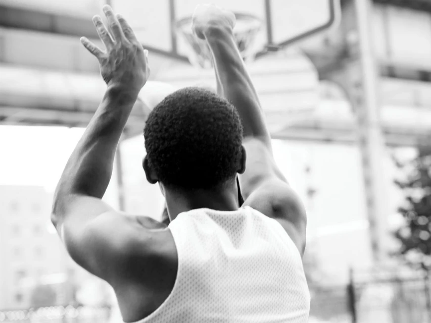 Black and white photo of an African American man in a white tank top shooting a basketball toward a hoop outdoors.
