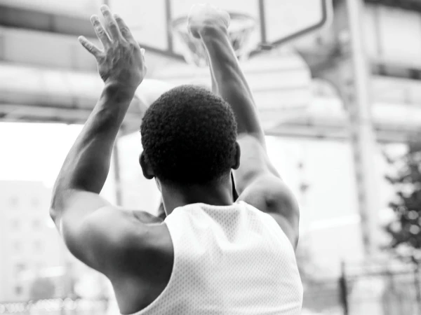 Black and white photo of an African American man in a white tank top shooting a basketball toward a hoop outdoors.