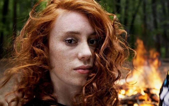 Close-up portrait of a young white woman with long curly red hair and freckles outdoors with a bonfire in the background.