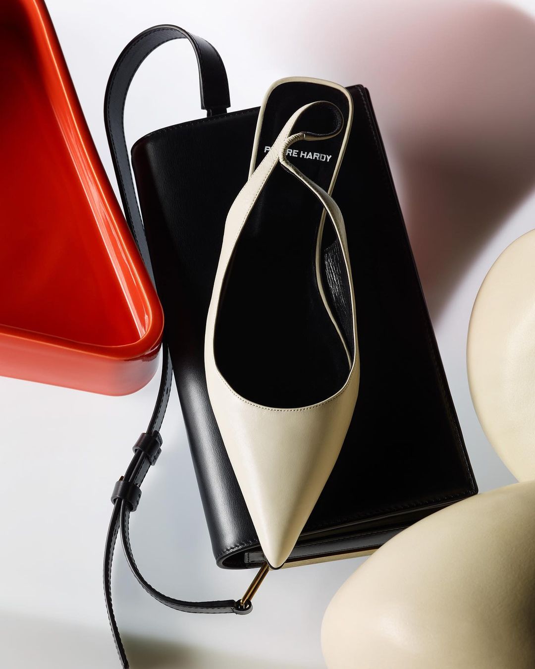 Fashion still life: cream-colored heeled mule on a black leather purse, with a red geometric dish and ivory sphere, studio lighting.