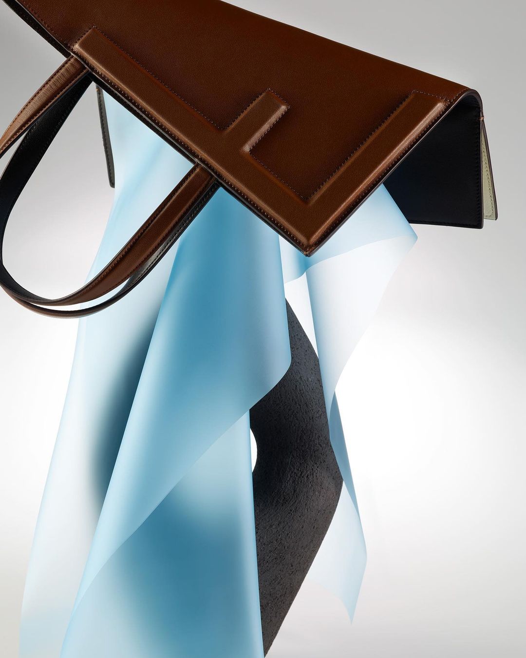 Elegant brown leather handbag with structured design, suspended against a soft blue fabric backdrop, creating a visually appealing contrast.