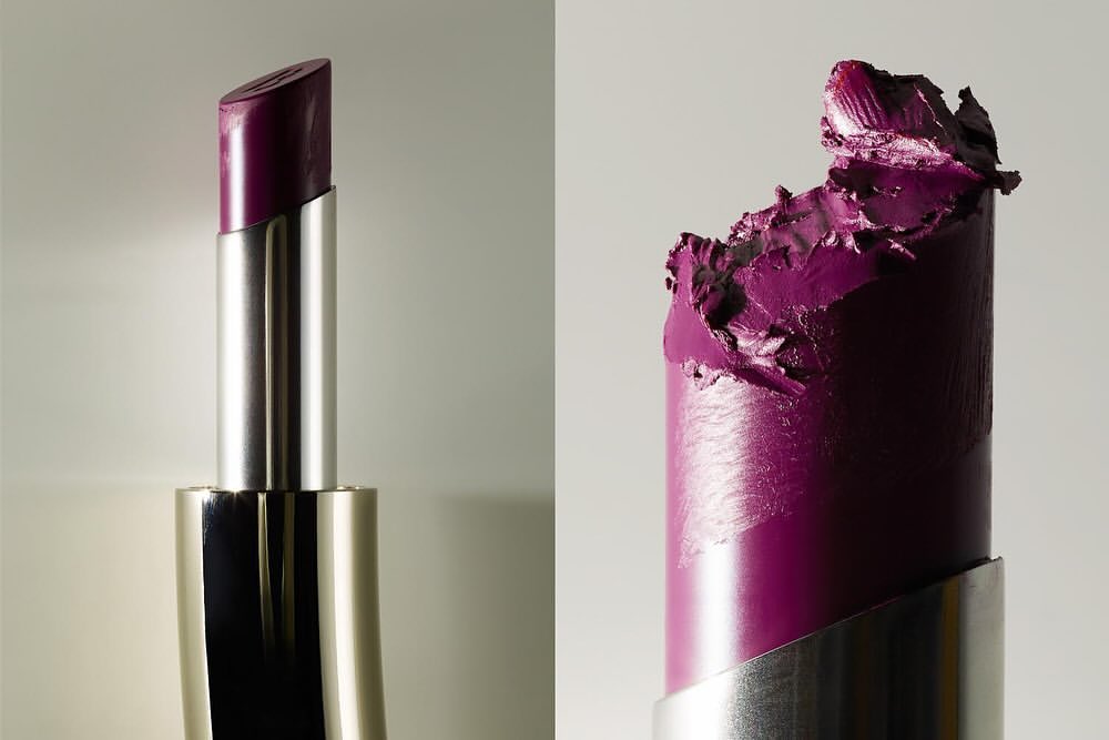 Two purple lipsticks, one pristine with a silver tube and one used with a silver tube on a neutral background. Beauty product shot.