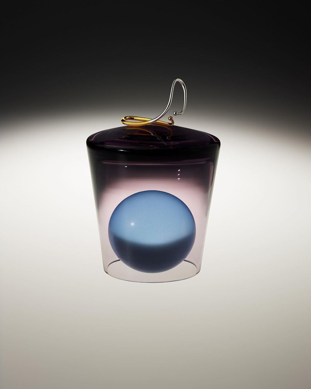 A clear glass container with a lavender-colored lid and handle holds a matte blue sphere, set against a dark gradient background.
