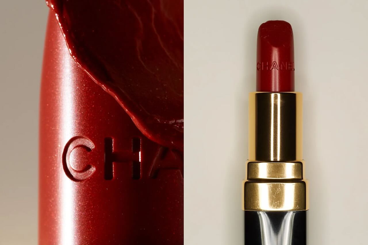 Close-up of a red Chanel lipstick, showcasing the product texture and logo detail. The cosmetic product is displayed against a neutral backdrop.