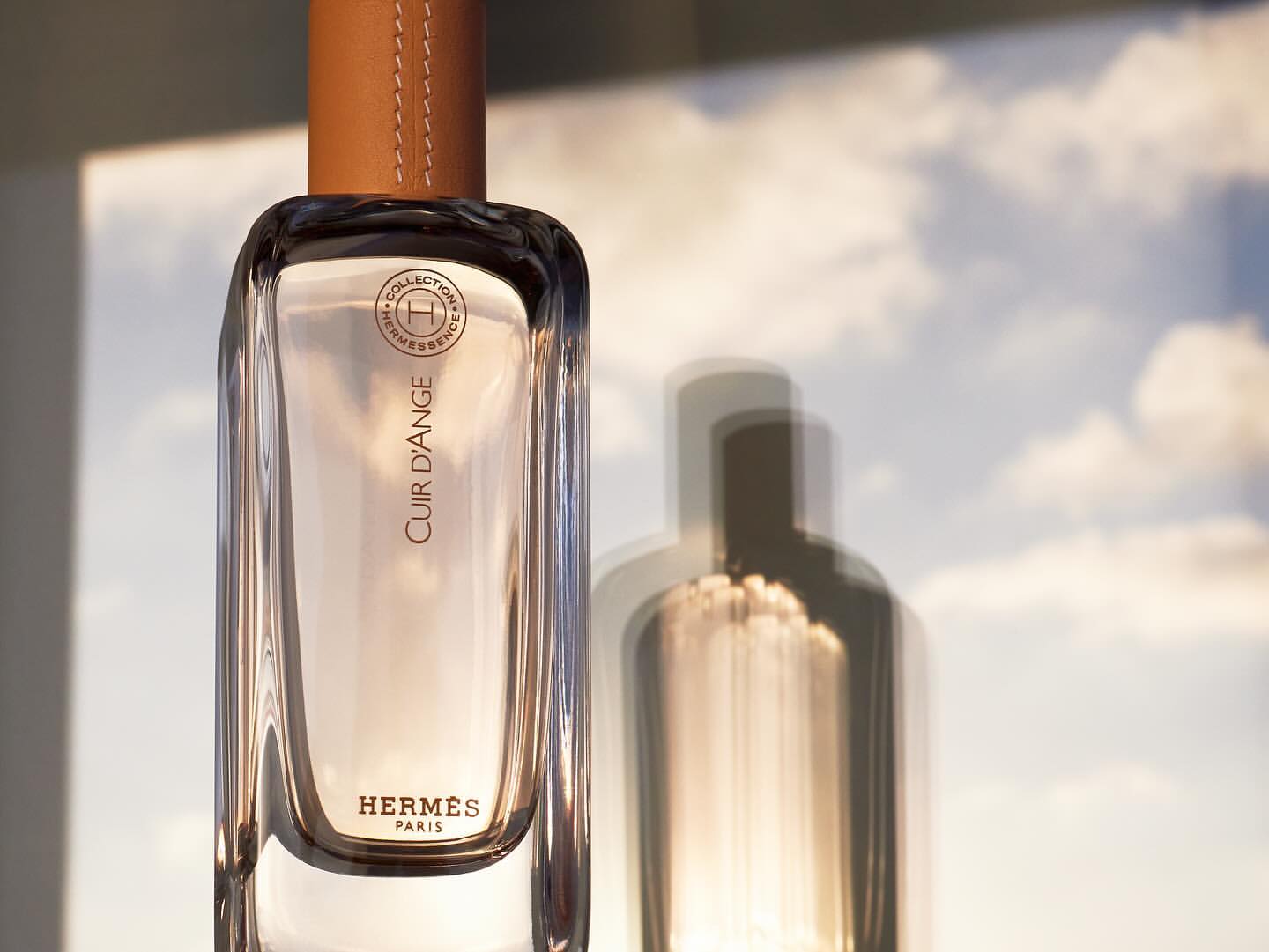 Product shot of Hermès Cuir d'Ange perfume bottle with leather top, set against a soft sky background, showcasing luxury and simplicity.