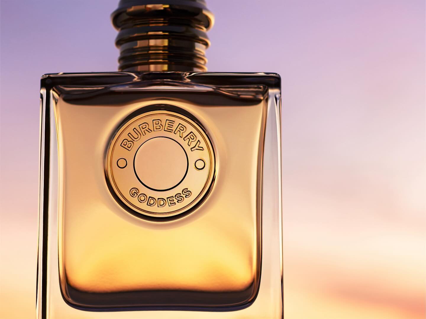 Burberry Goddess perfume bottle on a gradient purple and orange background. The glass bottle has a gold cap and a circular logo.