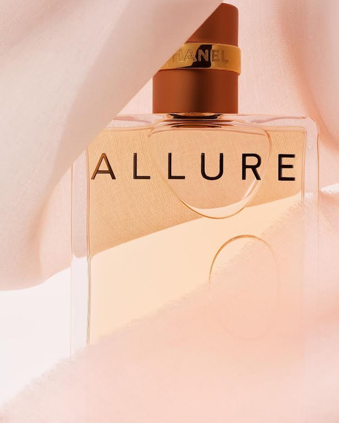 Close-up studio shot of Chanel Allure perfume bottle with gold accents, surrounded by soft pink fabric, against a pale pink backdrop.
