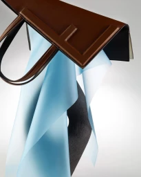 Structured brown leather tote bag with a folded blue paper sheet and dark sculptural form against a white background