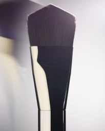 Close-up product shot of a makeup brush with dark dense bristles and a reflective handle against a neutral background.