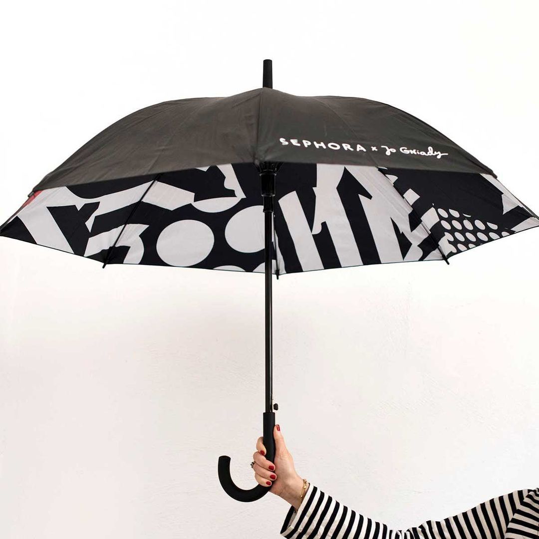 Person holding a Sephora x Joanie Charley black and white umbrella with a pattern of shapes against a white background.