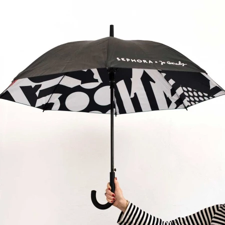 Person holding a Sephora x Joanie Charley black and white umbrella with a pattern of shapes against a white background.