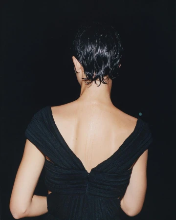 Back view of an adult woman with short, dark, wet hair wearing a black dress against a dark backdrop in an indoor setting.
