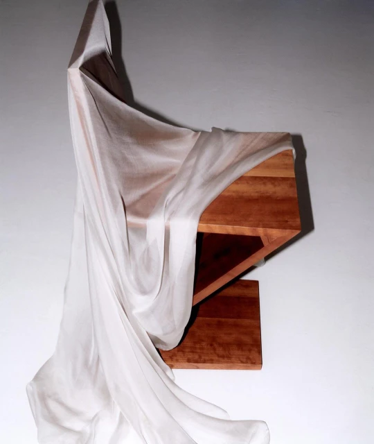 Wood shelf partially covered by a flowing white fabric against a neutral gray wall, creating a minimalist and modern aesthetic.
