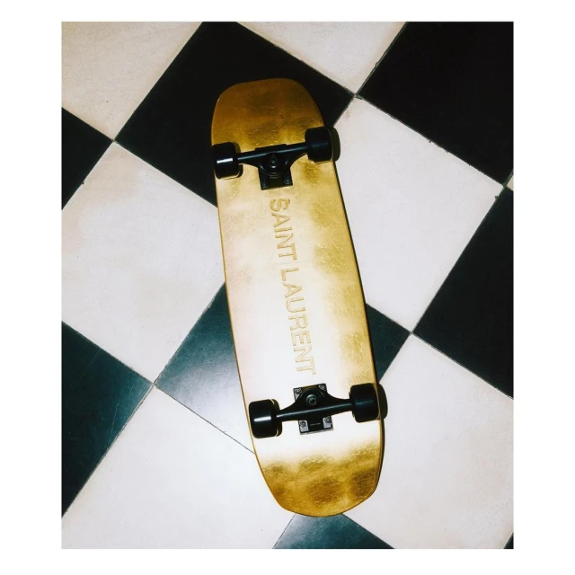 Gold Saint Laurent skateboard on a black and white checkered tiled floor. Fashionable skateboard with luxury brand visible in image.