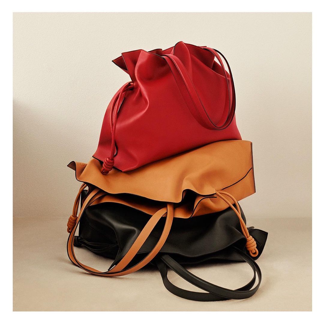 Stacked image of three luxury leather bags in red, tan, and black. The bags have a gathered top and drawstring closure. Studio lighting.