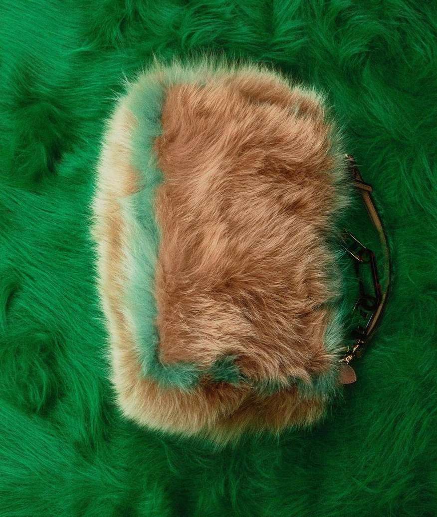Tan and turquoise faux fur purse with metal chain handle on a green faux fur background, studio shot.