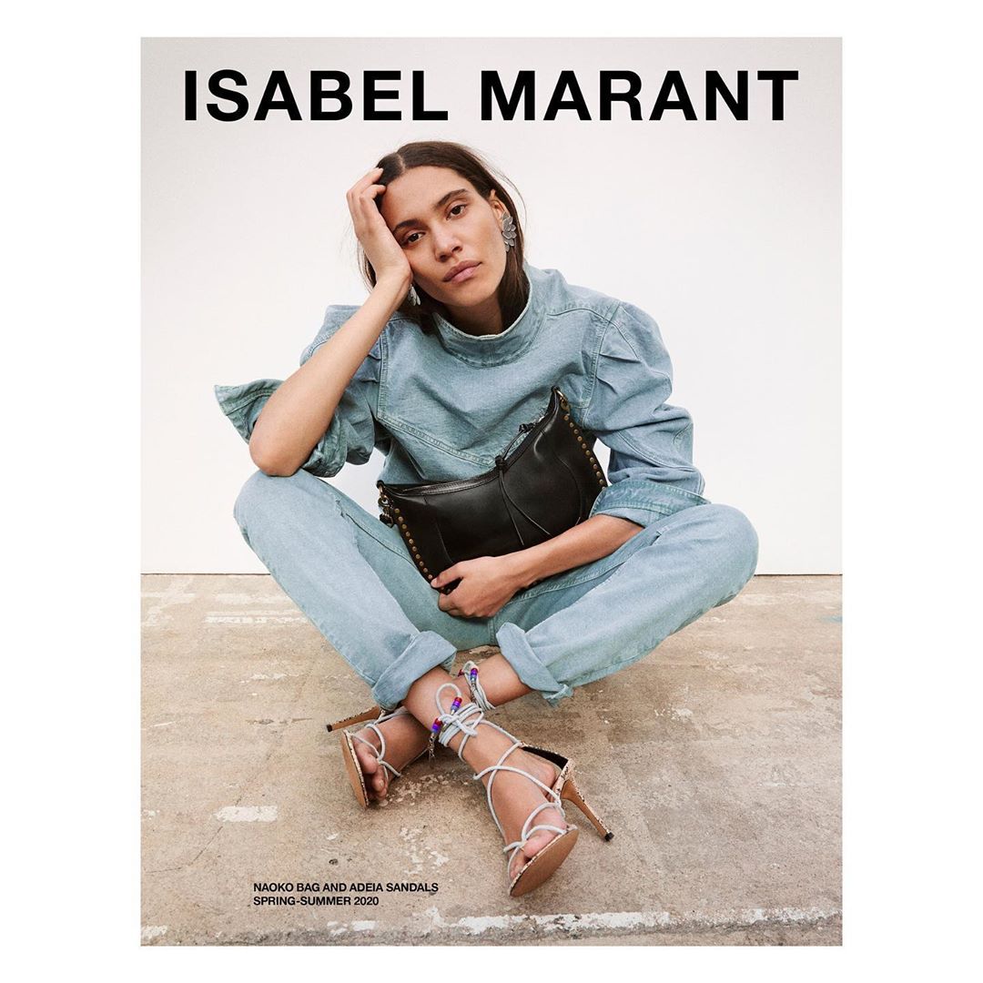 Fashion ad: Woman poses sitting in denim outfit with a black purse and strappy heels in a studio. Isabel Marant branding visible.