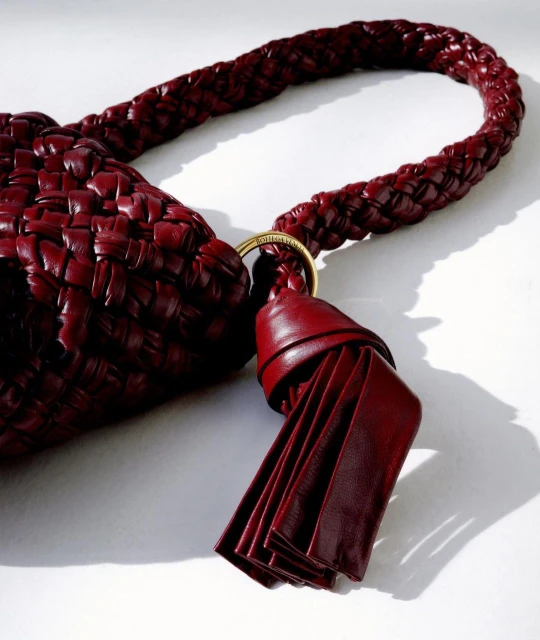 Close-up of a luxurious, woven red leather purse with a braided handle and a tassel, set against a simple, bright white backdrop.