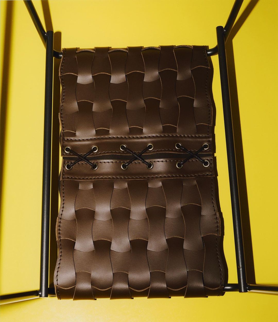 Brown woven leather chair supported by a black metal frame set against a bright yellow background in a studio.