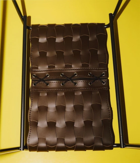 Brown woven leather chair supported by a black metal frame set against a bright yellow background in a studio.