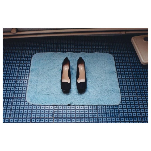 Overhead shot of black high-heeled shoes on a blue bathmat with a blue-tiled bathroom floor in the background in a domestic setting.