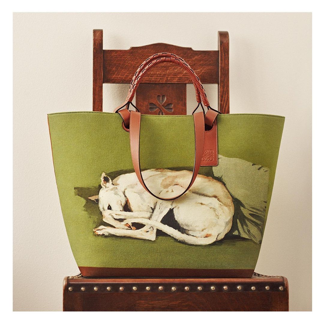 A green tote bag featuring a dog illustration is propped on a wooden chair, the bag has brown leather handles and accents, studio lighting.