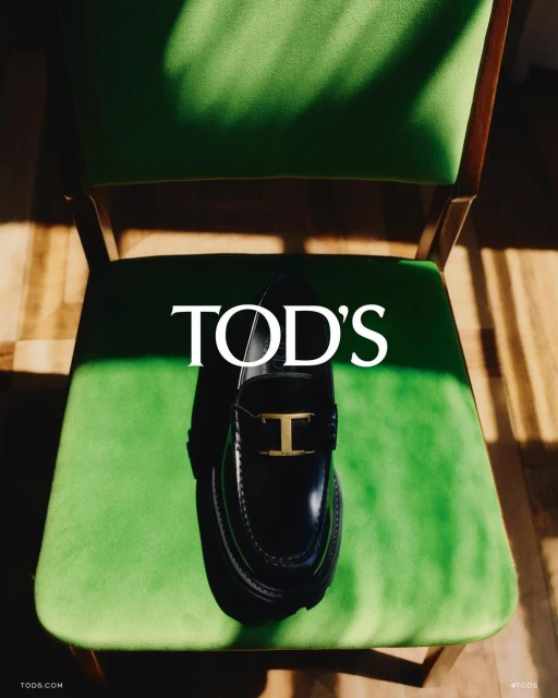 Tod's black leather loafer with gold buckle on green vintage chair, natural light and shadows. Fashion advertisement.
