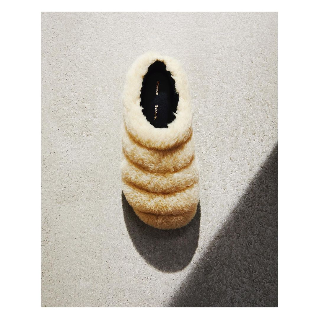 Beige, fluffy slipper with a dark insole is seen in a top-down shot on a textured gray surface with its shadow cast to the side.