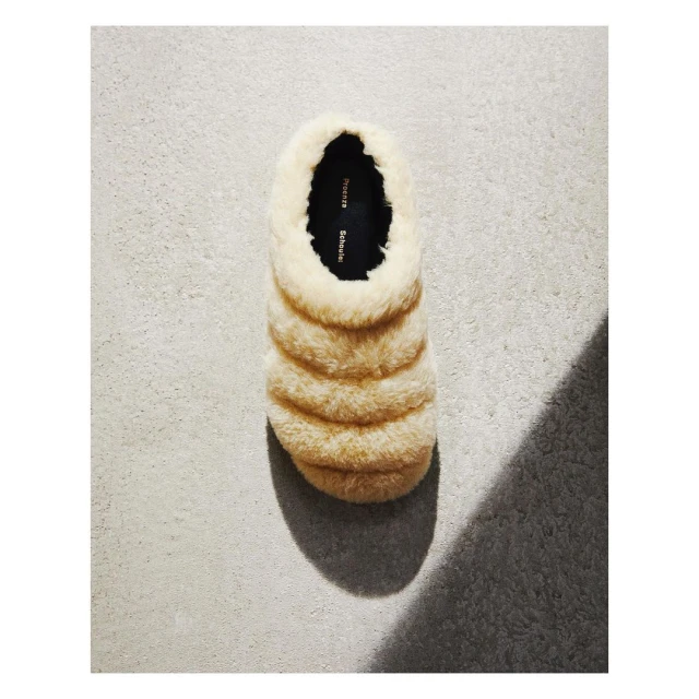 Beige, fluffy slipper with a dark insole is seen in a top-down shot on a textured gray surface with its shadow cast to the side.