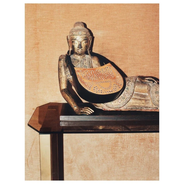 Buddha statue reclines with a studded leather purse. The statue sits on a modern table against a neutral textured backdrop.