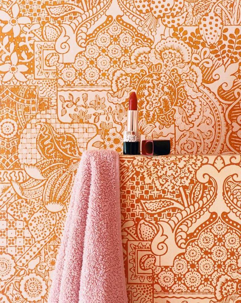 Close-up of a pink towel and red lipstick on a shelf against a patterned orange and white wallpaper in a bathroom setting.