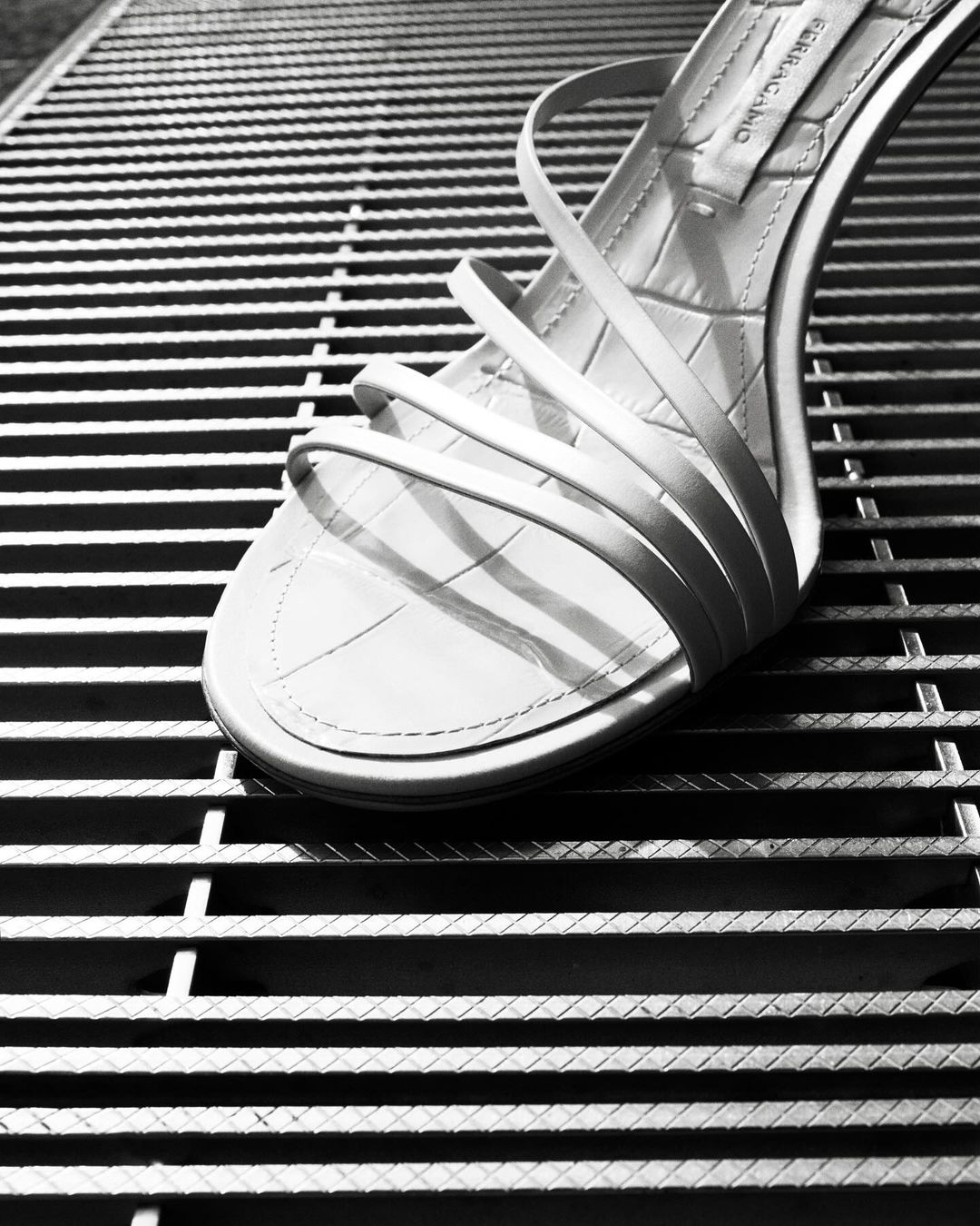 Elegant white strappy sandal on a textured metal grate. High heel shoe is the central focus in this minimalist fashion shot.