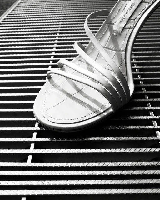 Elegant white strappy sandal on a textured metal grate. High heel shoe is the central focus in this minimalist fashion shot.