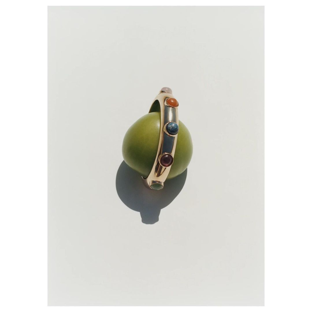 A gold ring with inset colored gemstones resting upright on a green olive against a clean white background.